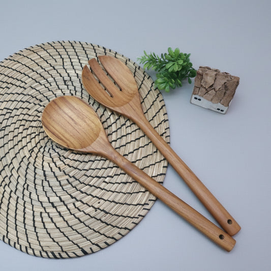 A pair of long-handled golden teak wood salad servers resting on a woven mat, showcasing natural wood grain.
