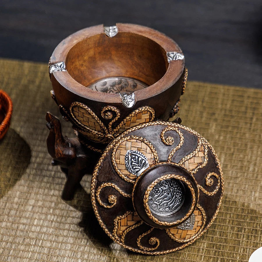 Vintage wooden ashtray with lid resting on three hand-carved elephant pillars, holding a cigarette.
