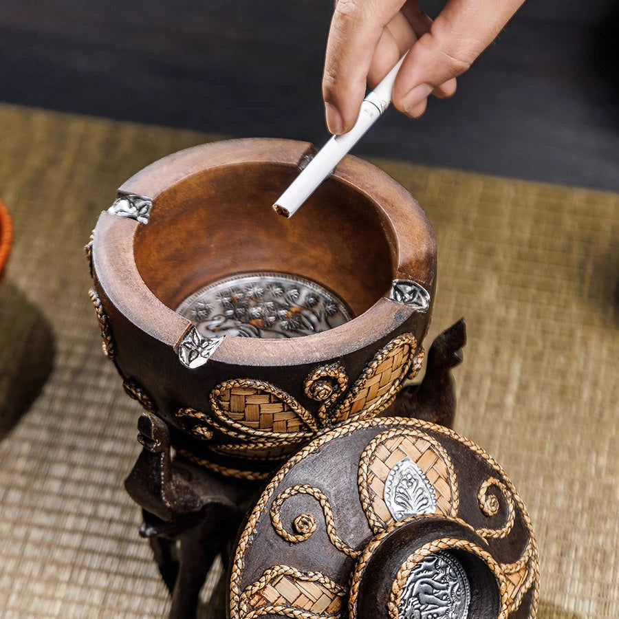 Vintage wooden ashtray with lid resting on three hand-carved elephant pillars, holding a cigarette.