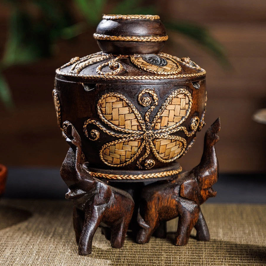 Vintage wooden ashtray with lid resting on three hand-carved elephant pillars, holding a cigarette.